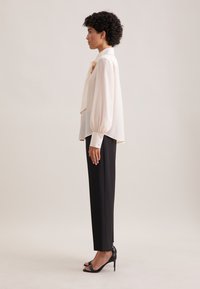 Cream blouse with puff sleeves and collar bow, paired with tailored black trousers. Model wears black heeled sandals. Smooth fabric texture.