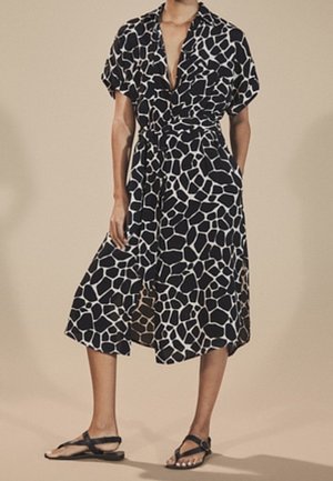 Frau trägt ein midi Kleid mit schwarz-weißem Giraffenmuster, kurzen Ärmeln und schwarzen flachen Sandalen, steht mit einer Hand in der Tasche.