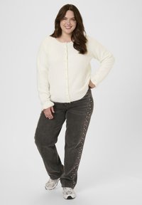 White fuzzy cardigan with buttons, paired with gray jeans featuring studded accents along the side. Model stands with a relaxed pose.