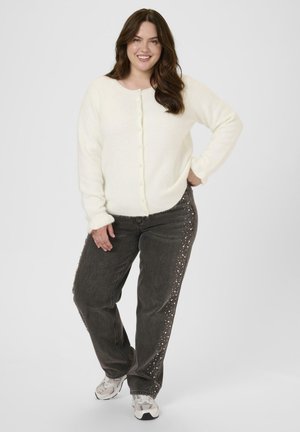 Weißer, flauschiger Cardigan mit Knöpfen, kombiniert mit grauen Jeans, die seitlich mit Nietenakzenten versehen sind. Das Model steht in einer entspannten Pose.