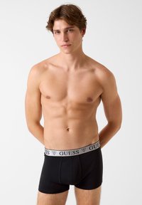 Čierne bavlnené boxerky s sivým elastickým pásom s logom "GUESS", ktoré nosí model na neutrálom pozadí.