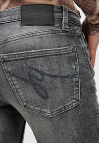 Κοντινό πλάνο πίσω τσέπης σε ξεθωριασμένο μαύρο τζιν με κεντημένο λογότυπο και μαύρο δερμάτινο ταμπελάκι επωνυμίας στη μέση.