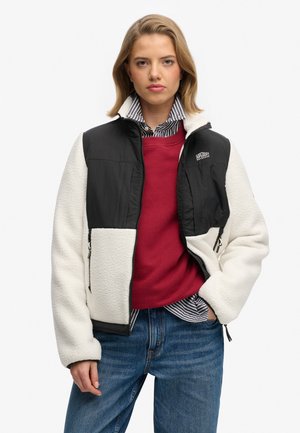 Jeune femme portant une veste polaire zippée noire et blanche par-dessus un pull rouge et une chemise rayée à col, jean bleu, debout avec une main dans la poche.