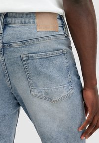 Vaqueros de mezclilla azul desgastados que cuentan con un bolsillo trasero y una etiqueta de cuero texturizado en color marrón claro con el logo "ALLSAINTS" grabado.