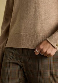 Beidzs krāsas kašmira džemperis ar ribotām apakšmalām un piedurknēm, savienots ar brūnām svītrainām biksēm, kas raksturo teksturētu audumu un vieglām krāsu variācijām.