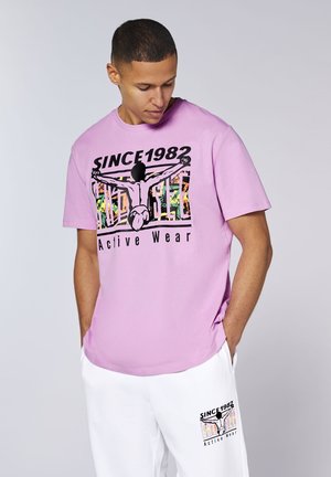 Mann trägt ein lavendelfarbenes T-Shirt und weiße Shorts mit dem Schriftzug „Since 1982 Active Wear“ und einer Grafik einer Person, die die Arme weit ausstreckt, auf beiden Kleidungsstücken.