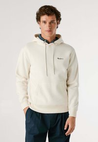Crème fleece hoodie met trekkoord, voorzak en geborduurd logo. Draagt over een donker shirt, met ribgebreide boorden en onderrand.