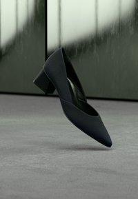 Crna cipela s niskom petom i špicastim vrhom lebdi iznad sive podloge, na zamućenoj sivoplavoj pozadini.