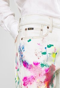 Bílé džíny z denimu mají vícolorý barevný rozstřik v růžové, zelené, modré a žluté. Obsahují logo "Polo", měděné kování a páskové smyčky.