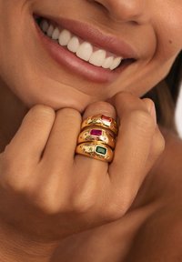 Persona sonriente sosteniendo el puño cerca de la cara, con tres anillos de oro que tienen piedras preciosas rojas, moradas y verdes, y grabados en forma de estrella.