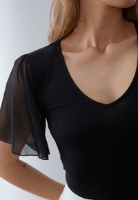 Černý top s přiléhavým střihem, s průsvitnými černými nafouklými rukávy a hlubokým výstřihem do V. Hladká textura s jemnými detaily.