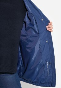 Interior de una chaqueta aislante azul marino, con forro suave y brillante, cierres a presión y una etiqueta de marca en la costura lateral.