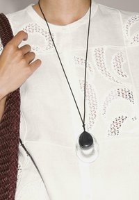 Black and silver pendant necklace on a black cord, worn over a white textured blouse with cut-out patterns. Burgundy woven bag held in hand.