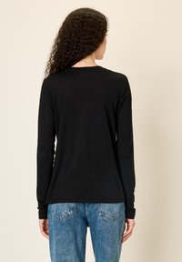 Pikkade lokkis juustega naine, kes kannab lihtsat musta pikkade varrukatega särk ja siniseid teksaseid, vaadatuna tagantpoolt beeži tausta ees.