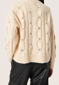 Suéter de punto en beige con patrón de trenzas, que presenta detalles calados, un dobladillo acanalado y un ajuste relajado. Llevado sobre pantalones de cuero negros.