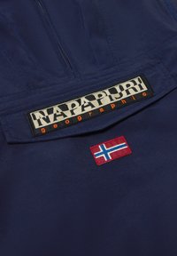 Marineblå jakke med høy krage og glidelås, et fremtredende "Napapijri" logo-patch og et brodert norsk flagg. Jevn tekstur.
