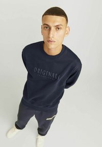 Marineblauwe sweatshirt met gestructureerd "ORIGINALS" logo en bijpassende broek. Glad materiaal, ronde halsontwerp, gecombineerd met lichtgekleurde sneakers.