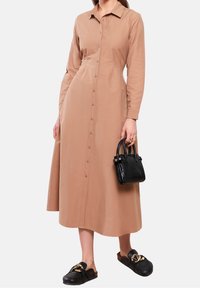 Abito lungo con bottoni in tessuto beige, dotato di colletto e maniche lunghe. Abbinato a una borsa nera con stampa coccodrillo e scarpe nere slip-on.