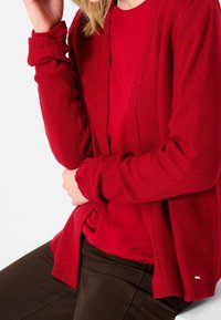 Cardigan rosso con texture, dalla vestibilità rilassata, indossato sopra una maglietta rossa coordinata. Presenta polsini a coste e un piccolo logo decorativo. Pantaloni dalle tonalità terrose.
