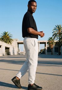Mann in schwarzem T-Shirt, hellgrauer Jogginghose und schwarzen Sneakers steht an einem sonnigen Tag draußen mit Palmen und modernen Gebäuden im Hintergrund.