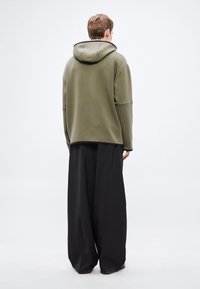 Olivovo zelená mikina s kapucňou a voľným strihom, nosená cez čierne tričko. Tkanina je hladká s jemným leskom a dlhými rukávmi. Široké čierne nohavice.