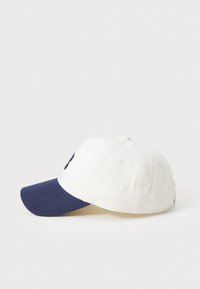 Bílá baseballová kšiltovka s námořnickým modrým kšiltem, na přední straně s námořnickým logem. Vyrobena z bavlny s pevným designem.