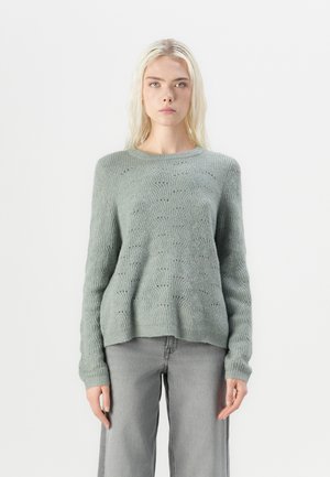Un pull en maille vert clair avec un motif texturé, un col rond et des manches longues. Associé à un pantalon large gris.