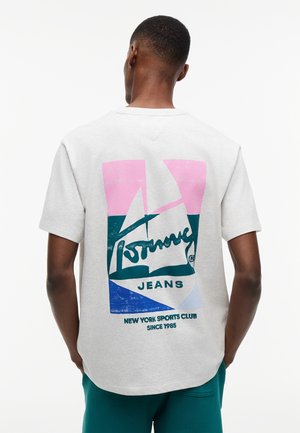 Mann trägt weißes T-Shirt mit kurzen Ärmeln, farbenfrohem geometrischem Muster und dem Text "Tommy Jeans" sowie "New York Sports Club Since 1985" auf dem Rücken.