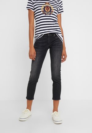 Person iført hvide sneakers, cropped mørke vaske smalle jeans og en navy og hvid stribet skjorte med et våbenskjold logo.