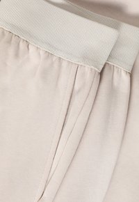 Pantaloni de trening de culoare bej deschis, cu un tiv elastic cu textură pliată, o textură moale și buzunare laterale, proiectați pentru confort și ușurință în mișcare.