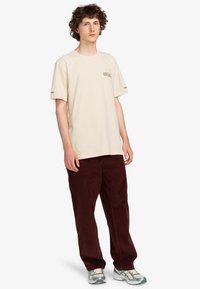 Beige katoenen t-shirt met korte mouwen en een bedrukt logo, gecombineerd met bordeauxkleurige corduroypants en grijze sportschoenen.