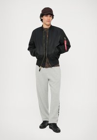Jachetă neagră tip bomber cu fermoar aurie, pantaloni de trening gri cu text pe lateral și adidași negri. Prezintă o textură detaliată a materialului și o croială casuală.