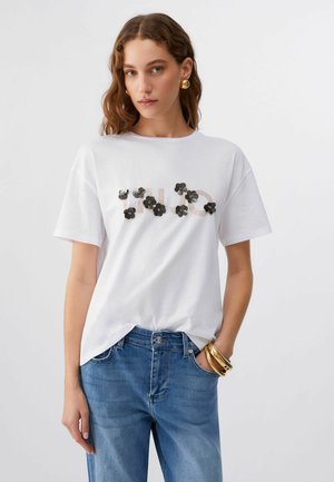 Donna con capelli castani mossi indossa una T-shirt bianca decorata con applicazioni di fiori neri sopra un testo, abbinata a jeans blu e gioielli dorati.