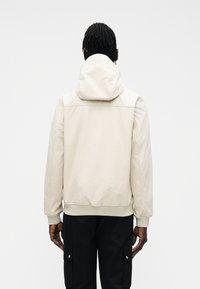 Gaiši bēša kapuce ar mīkstu tekstūru, regulējamu kapuci un ribotu apakšu. Vienkārša dizaina, redzama no aizmugures.