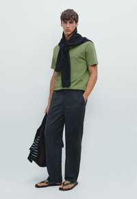 Groen T-shirt met korte mouwen, zwart geribd vest over de nek gedrapeerd, donkere broek, zwarte slippers, draagt een donkere tas met een gestreept patroon.