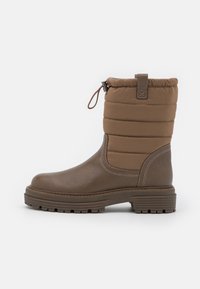 Botte imperméable marron avec un dessus en tissu matelassé rembourré, semelle en caoutchouc et bout arrondi. Elle dispose d'une languette et d'un cordon élastique en haut.