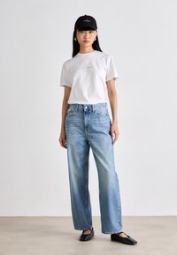 Hvit bomulls t-skjorte med en liten logo, kombinert med høyhåndet blå denim vidbukser. Svart flate sko og en svart caps fullfører antrekket.