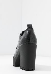 Zapato de tacón negro con un upper de cuero texturizado con patrón de cocodrilo y un tacón sólido negro y grueso. Presenta una lengüeta trasera para facilitar el calzado.