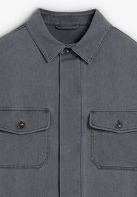 Camisa de denim gris con cuello punta, dos bolsillos en el pecho con cierres de solapa y detalles de botones. Tejido texturizado con un diseño estructurado.