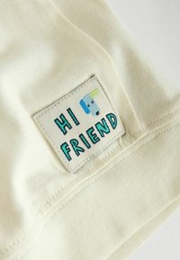 Tissu de couleur crème avec une étiquette cousue portant l'inscription « HI FRIEND » en vert, et une illustration de chien en bleu et jaune. Texture douce.