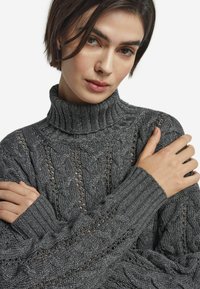 Grau gestrickter Pullover mit hohem Rippenkragen, strukturierten Zopfmuster und glitzerndem Finish. Mit Rippenbündchen und Nähdetails.