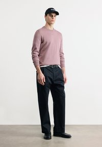 Νέος άνδρας που φοράει ροζ πουλόβερ με σκονισμένη απόχρωση, μαύρο παντελόνι φαρδιάς γραμμής, μαύρη ζώνη, μαύρα παπούτσια και μαύρο καπέλο με άσπρο κείμενο, στέκεται σε γκρι δάπεδο.