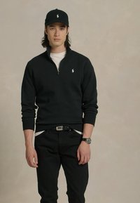 Zwarte rits sweatshirt met een klein logo, gecombineerd met een zwarte pet en strakke zwarte broeken. Een horloge met een ronde wijzerplaat is zichtbaar om de pols.