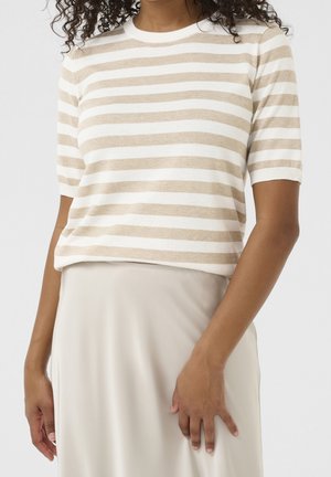 Femme portant un haut à manches courtes beige et blanc à rayures horizontales, rentré dans une jupe crème taille haute, sur un fond uni.
