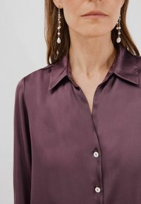 Blusa di raso viola con scollatura colletto, chiusura con bottoni e una leggera lucentezza. Orecchini pendenti con perle e dettagli in metallo.