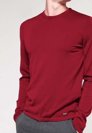 Mann trägt einen eng anliegenden roten Pullover mit langen Ärmeln, in graue Hosen gesteckt, Hände in den Taschen, Ausschnitt oberhalb des Kinns.