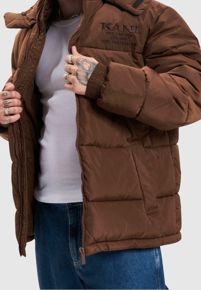 Brun puffjacka med dragkedja fram, ribbade ärmslut och sidofickor. "KANI"-logotyp broderad på bröstet, med en mjuk och glänsande yta.
