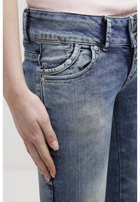 Close-up of a person’s left hand resting on blue faded denim jeans with visible front pocket stitching and metal rivets.
