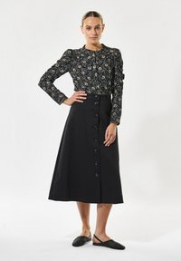 Tamna cvjetna bluza s dugim rukavima, crna suknja s prednjim gumbima i džepovima, te crne perforirane slip-on cipele.