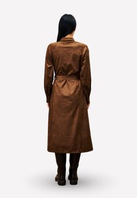 Frau mit langen schwarzen Haaren, die einen braunen Mantel mit Gürtel und schwarze Stiefel trägt, steht mit dem Rücken zu einer schlichten Hintergrund.